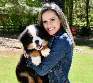 Riggie The Berner at 12 weeks old being held by his mommy, Katie, and they are outside standing on grass with a wooded background.