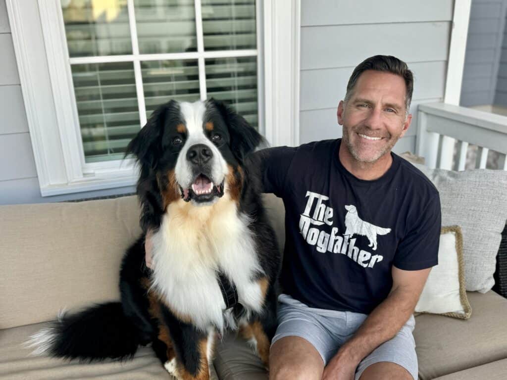 Riggie the Berner sitting on a couch with his Dad, who is wearing a t-shirt that says "The Dogfather".