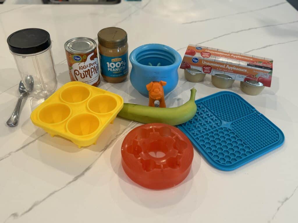 An assortment of food-focused canine enrichment tools including Pupsicles, a Toppl, and a lick mat along with pumpkin, apple sauce, peanut butter, and a banana.