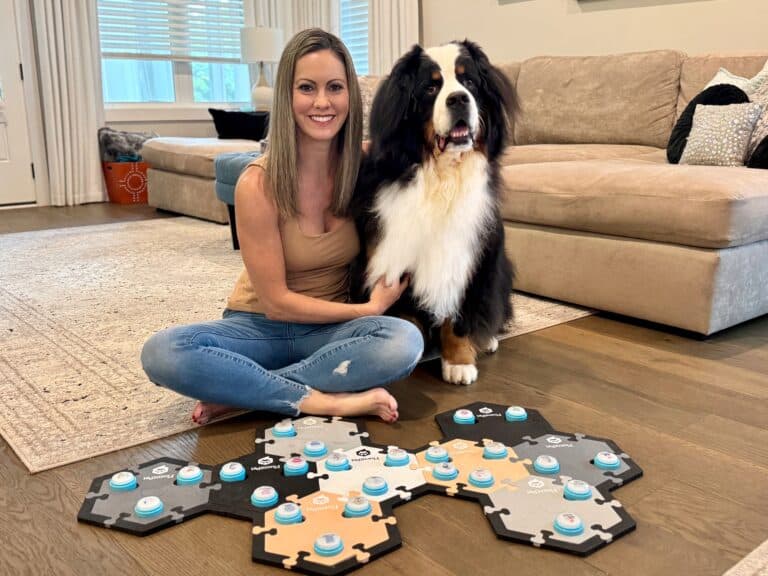 A woman with light brown hair sitting on the floor with her arm around a Bernese Mountain Dog with a FluentPet soundboard with dozens of dog buttons in front of them.