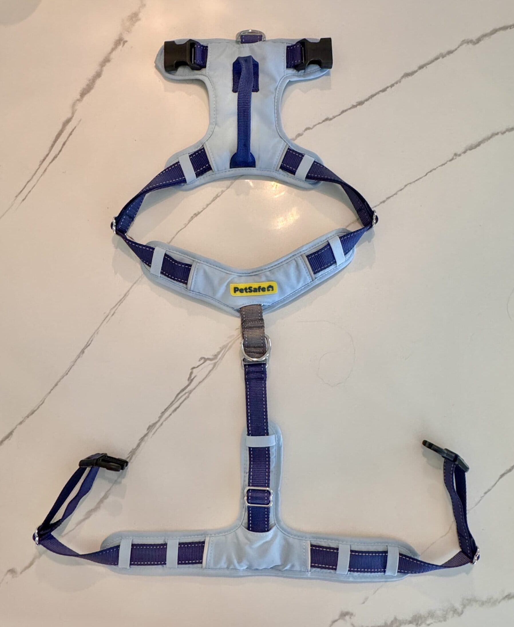 A photograph of an unhooked PetSafe sky blue dog harness with royal blue straps and a handle lying flat on a white marble surface.